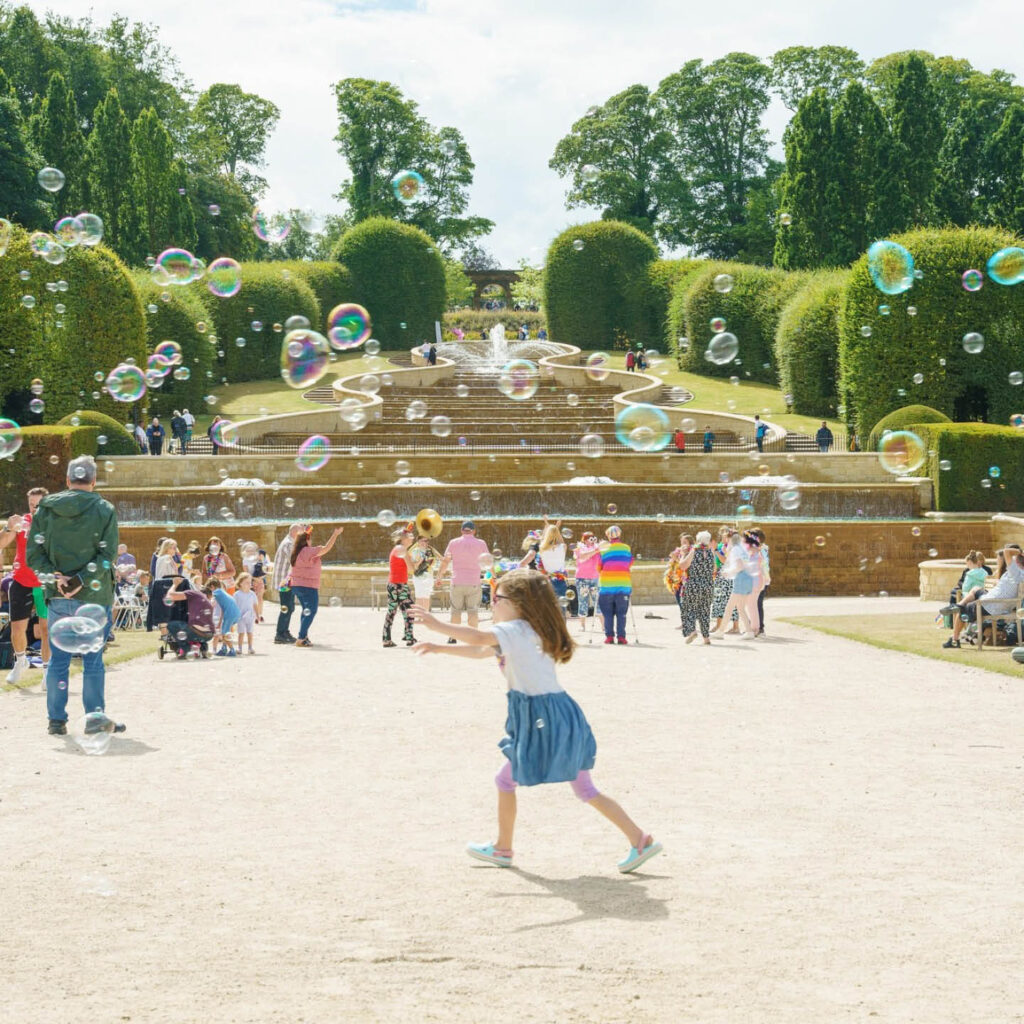 Children play in the sun at the bottom of a cascading fountain at Alnwick Garden. Big iridescent bubbles float above and around them.