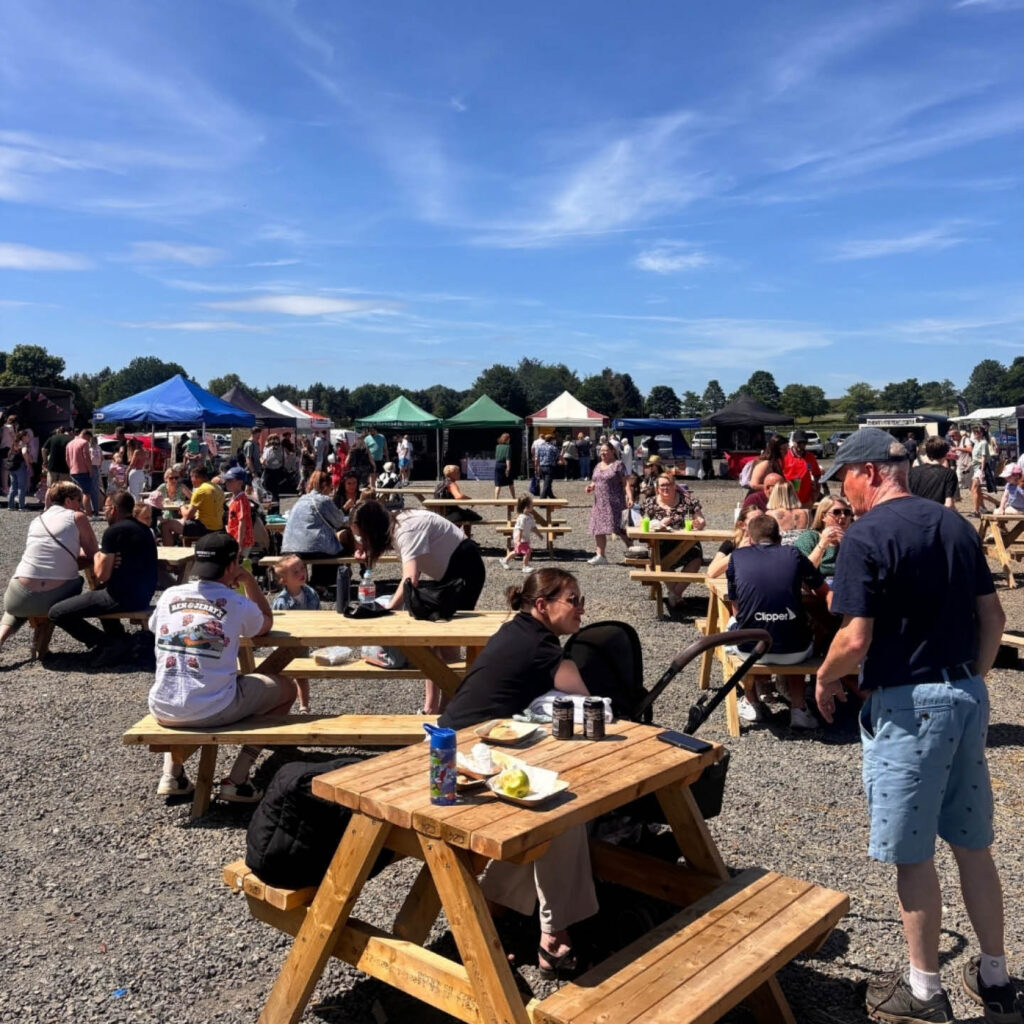 People sit at picnic benches underneath a blue summer sky at Brocksbushes Farm. Markets stalls can be seen in the distance.