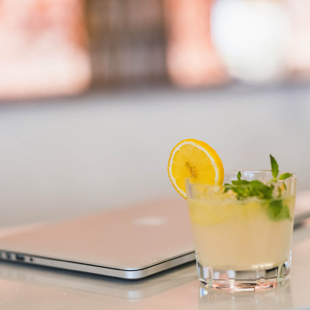 A closed laptop on a white tabletop. A tumbler containing a cloudy cocktail with a lemon slice sits next to it.