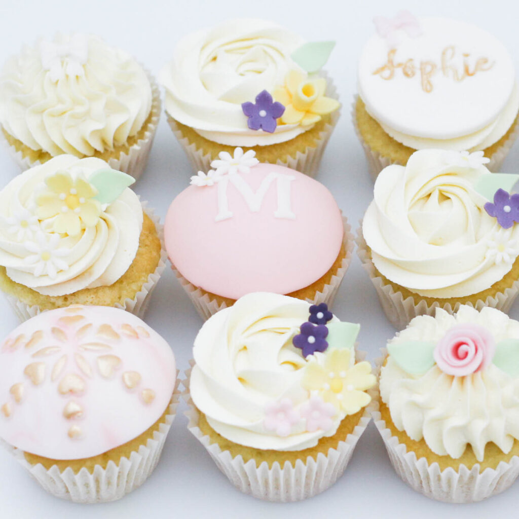 Beautiful cupcakes topped with pale pink and white frosting. They are decorated with dainty flowers, lettering and metallic patterns.
