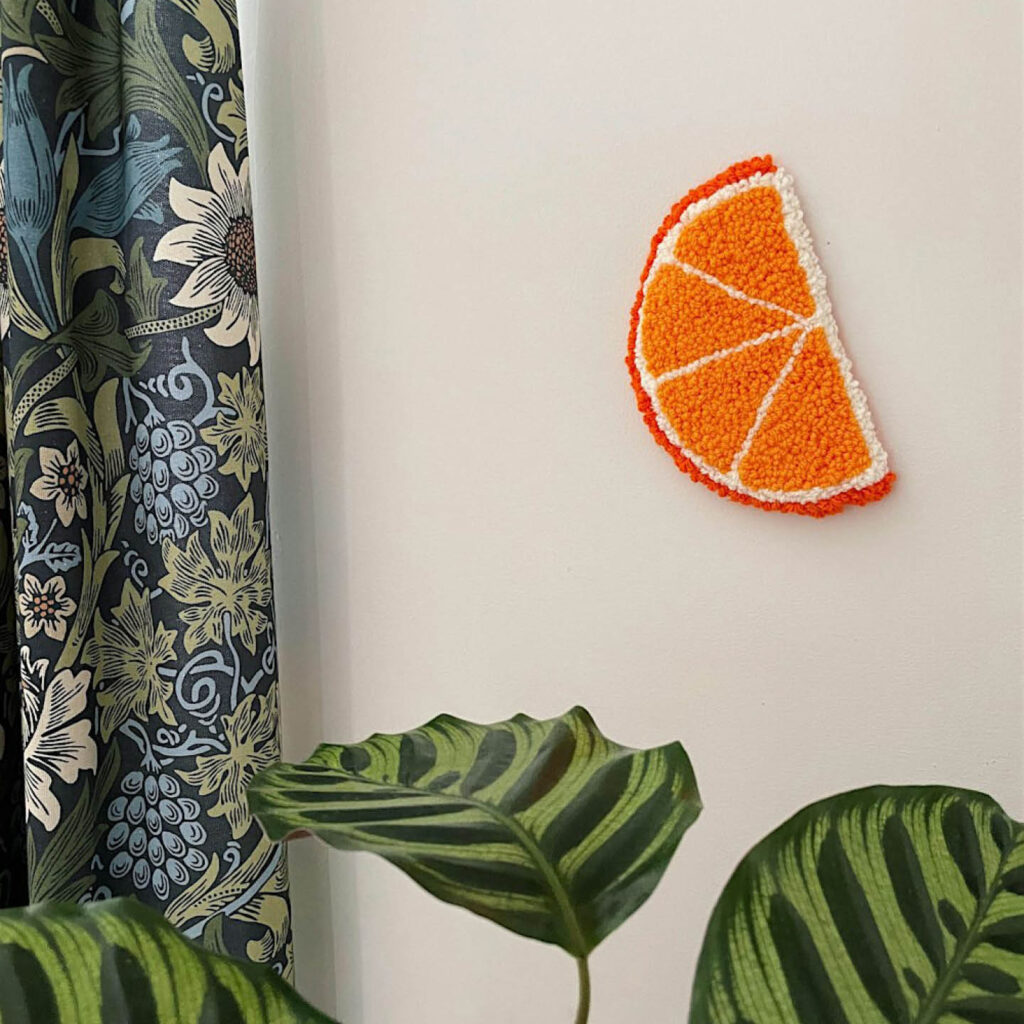A punch needled orange slice hangs from a white wall above a large plant. It is one of the hangings you can make at the fruit punch needling workshop at Flaura Rose.