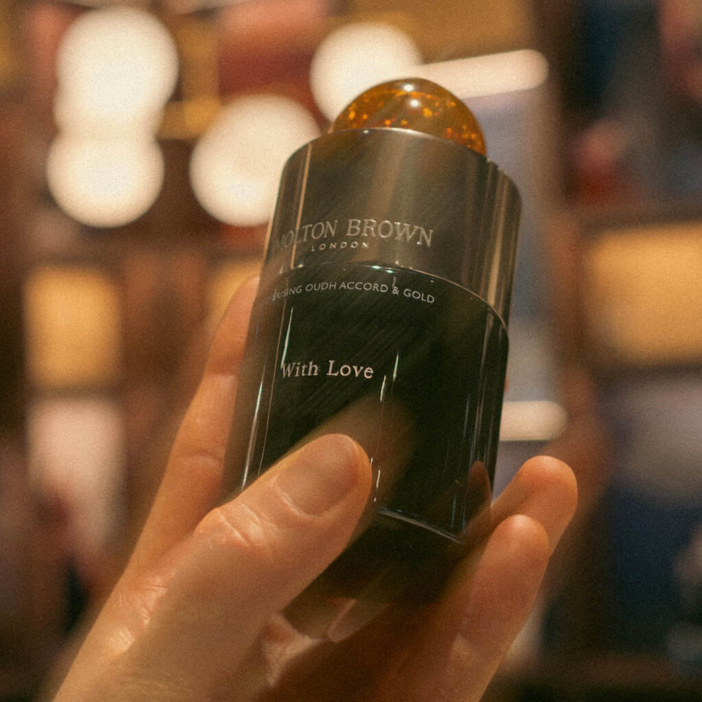 A hand holds up a black Molton Brown fragrance bottle with an amber coloured circular stopper in the top. White text printed across it says: Molton Brown, Mesmerising Oudh Accord & Gold.