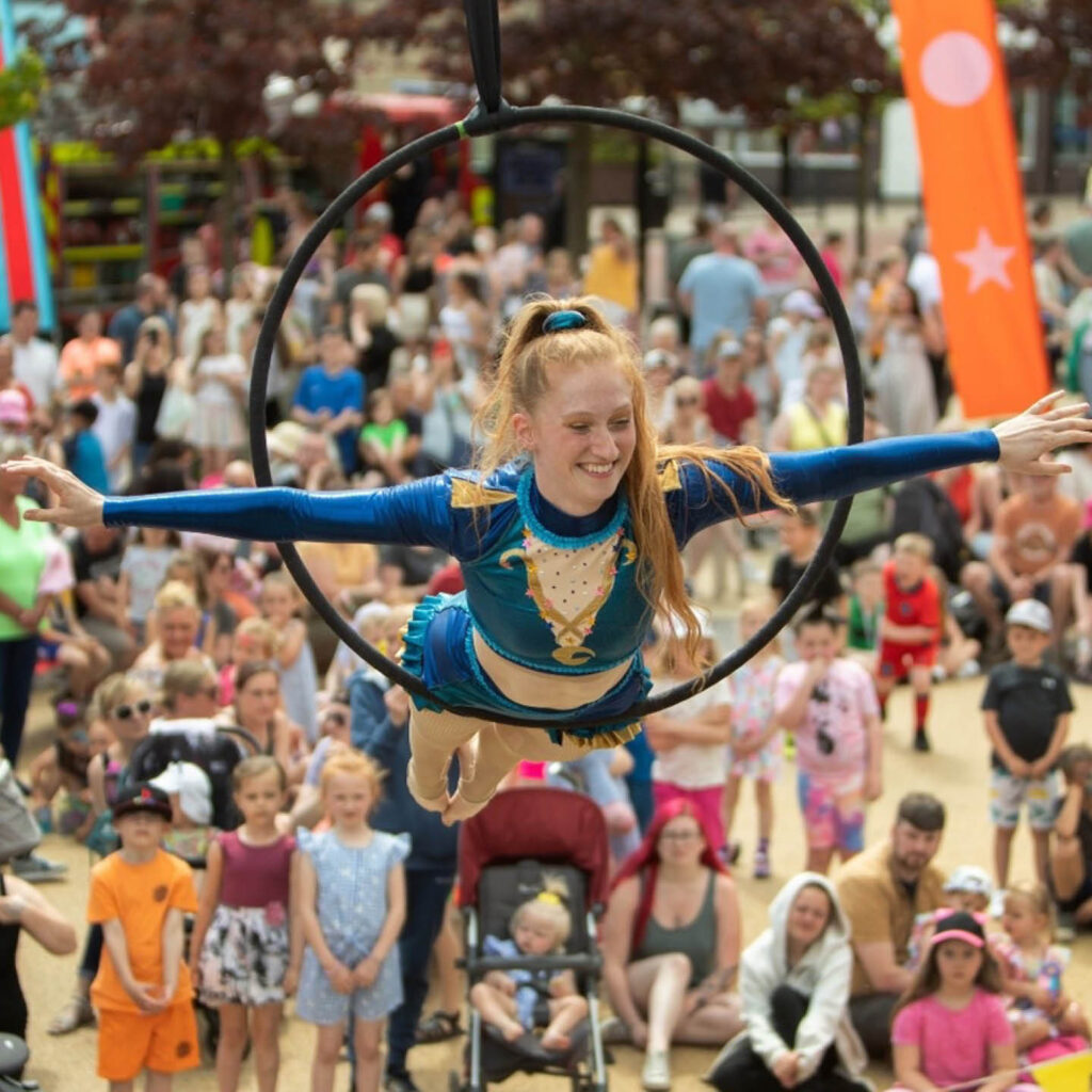 A young woman wearing a blue circus leotard grins as she balances her stomach on an aerial hoop. A crowd gathers below to watch the show. She is performing at Newcastle Quayside's carnival.
