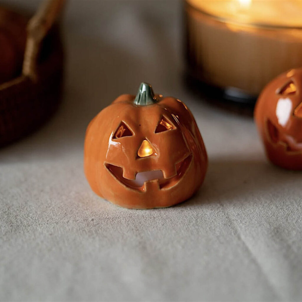 A smiling pumpkin tealight holder on a white cloth surface. A candle burns inside it.