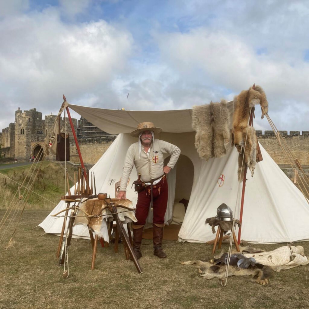 A man dressed in white medieval attire stands proudly in front of a white tent draped with animal skins and furs. He is one of the actors at the Rosa Mundi encampment at Alnwick Castle.