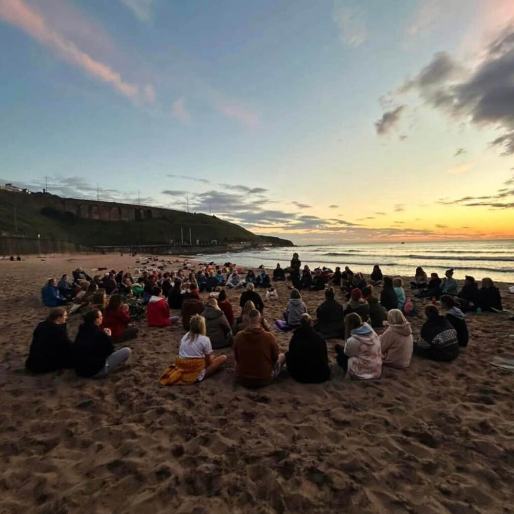A big group of people sit on the sand of King Edward's Bay, Tynemouth. The sky is streaked yellow and pink with the sunrise. They are taking part in the Shore Yoga autumn equinox session.