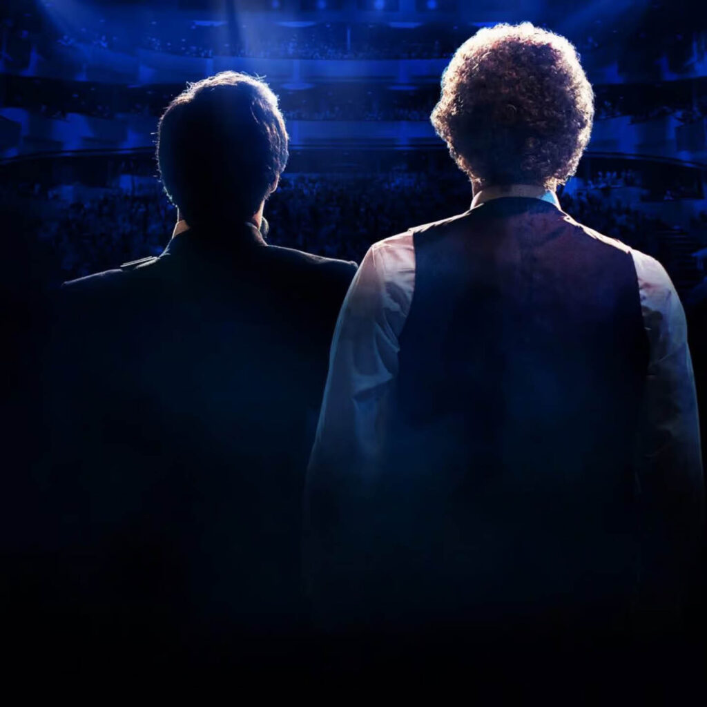 Two men stand next to each other and look out into the darkness of a stadium. They are cast in dark blue shadow. This is the poster for The Simon and Garfunkel Story.