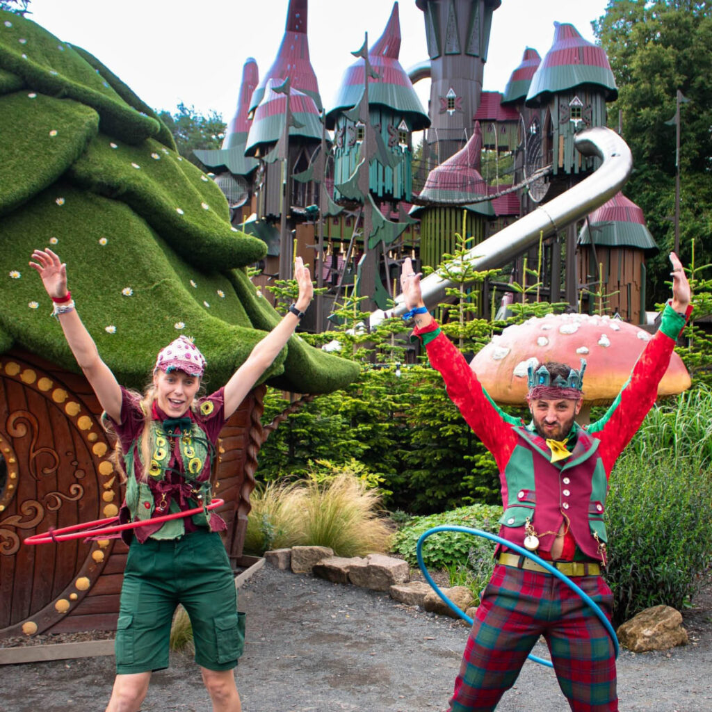 Two people dressed in elf like costumes raise their arms in the air and pull funny faces as they hula hoop. They stand between little grass houses. Behind them is a fairytale castle structure. They are two of the cast members during Spritember at Lilidorei, in Alnwick Garden.