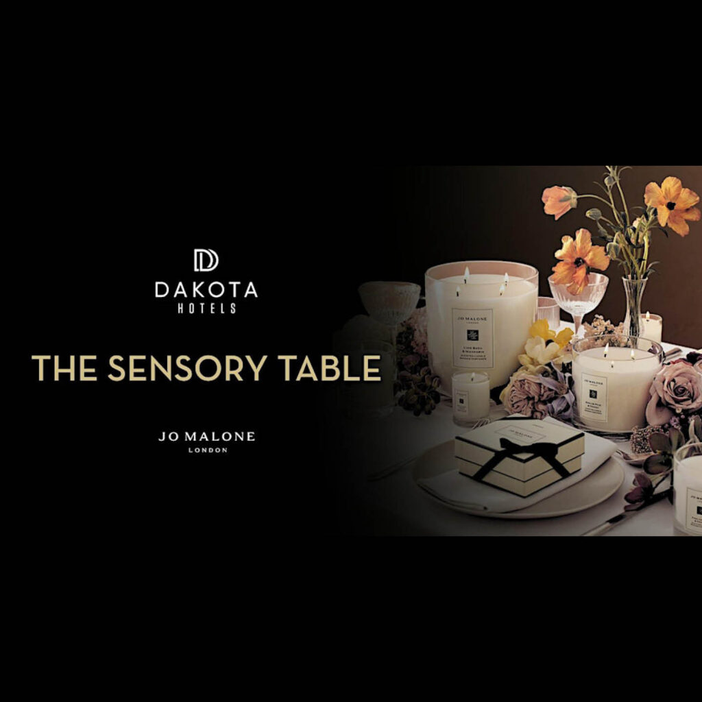 Jo Maline candles decorate a table alongside a vase of orange flowers, plates, and letters tied with black ribbon. White text against a black background reads: Dakota Hotels, The Sensory Table, Jo Malone.