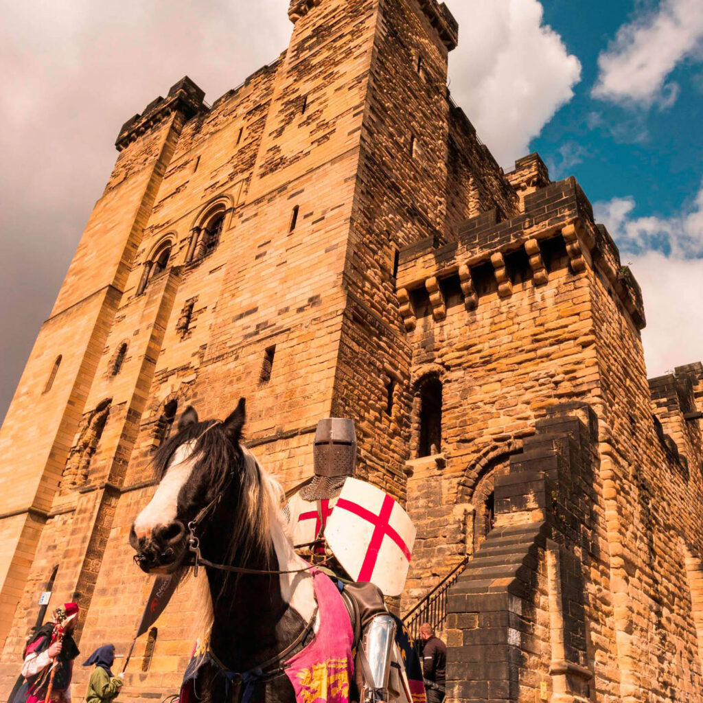 A knight with a shield painted with the England flag sits on the back of a brown horse. Newcastle Castle looms behind them in the sun.