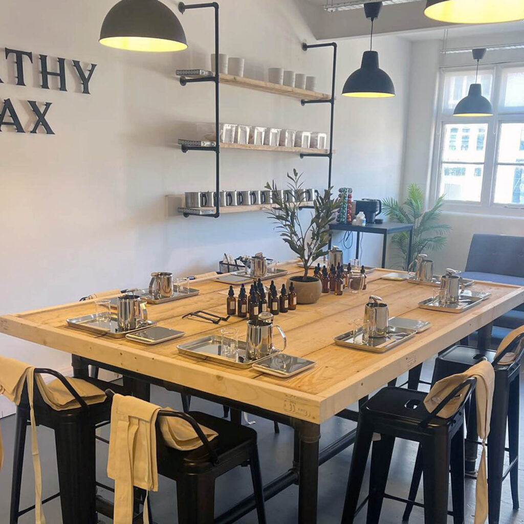 A wooden table with black stools draped with white aprons. The table is laid with candle making equipment and fragrance bottles. This is the Worthy Wax studio prepared for a candle making workshop.