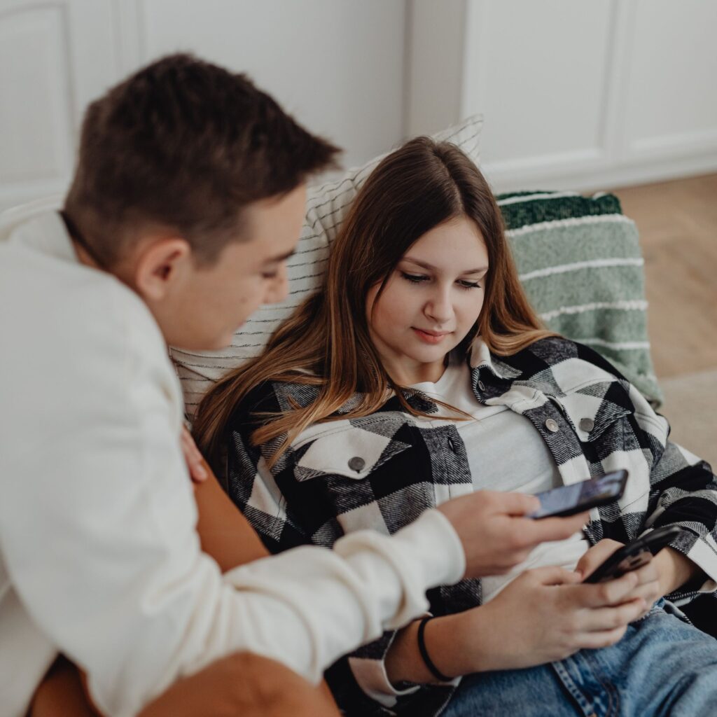 Two teenagers are sitting on a couch showing each other their phone screens