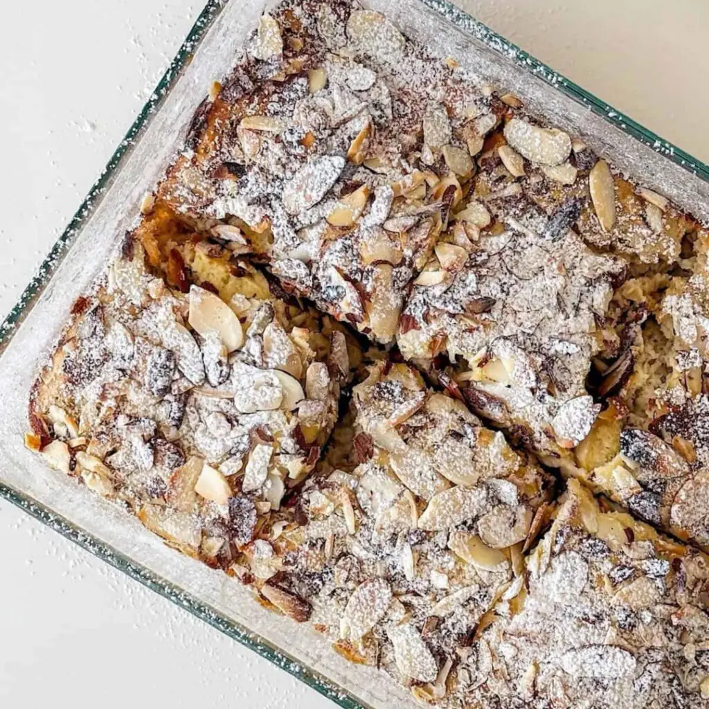 A bowl of baked almond oats topped with flaked almonds and a drizzle of honey, served warm with a spoon. The golden-brown oat bake has a soft, fluffy texture and is garnished with fresh berries for a wholesome breakfast.
