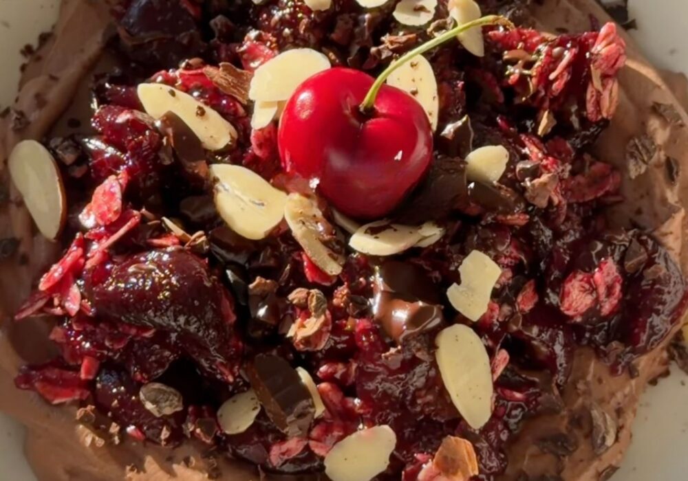 A bowl of creamy chocolate yogurt topped with a warm cherry oat crumble made with juicy cherries, rolled oats, maple syrup, and cinnamon, finished with sliced almonds, cacao nibs, chopped dark chocolate, and a fresh cherry.