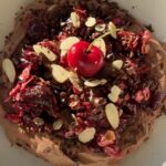 A bowl of creamy chocolate yogurt topped with a warm cherry oat crumble made with juicy cherries, rolled oats, maple syrup, and cinnamon, finished with sliced almonds, cacao nibs, chopped dark chocolate, and a fresh cherry.