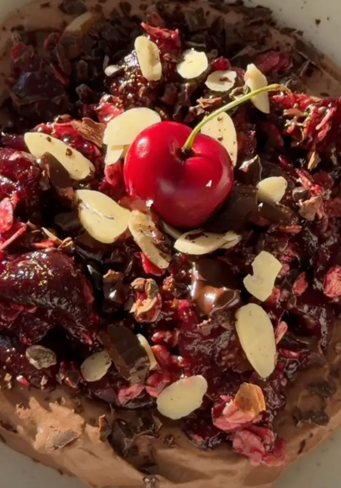 A bowl of creamy chocolate yogurt topped with a warm cherry oat crumble made with juicy cherries, rolled oats, maple syrup, and cinnamon, finished with sliced almonds, cacao nibs, chopped dark chocolate, and a fresh cherry.