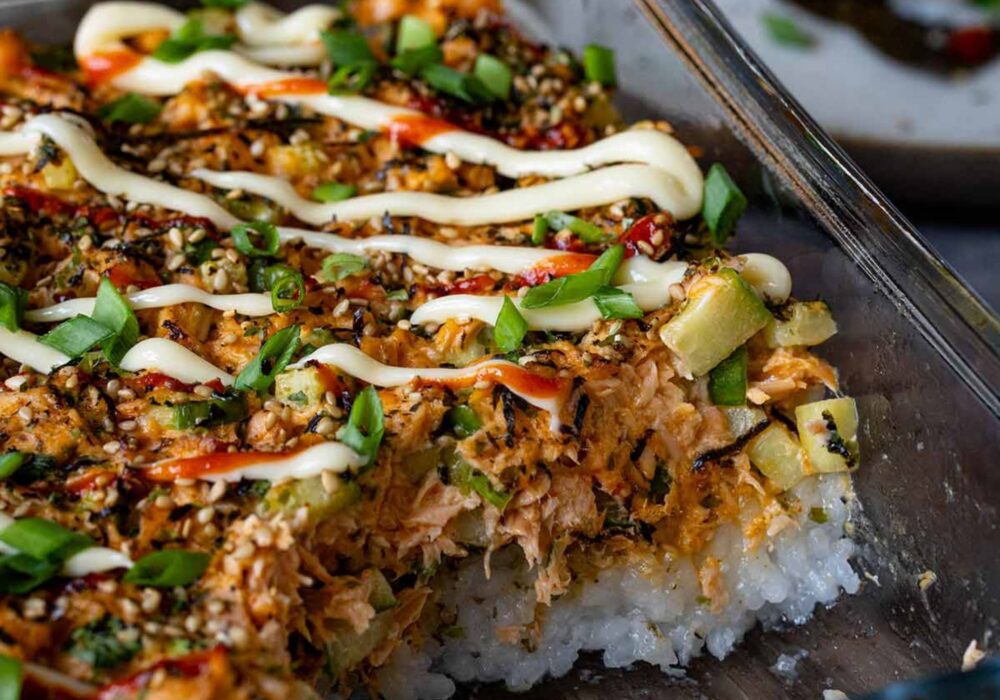 A close-up of a baked salmon sushi dish topped with sesame seeds, spring onions, spicy sauce and creamy mayonnaise, served in a glass baking dish.