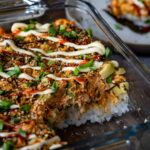 A close-up of a baked salmon sushi dish topped with sesame seeds, spring onions, spicy sauce and creamy mayonnaise, served in a glass baking dish.