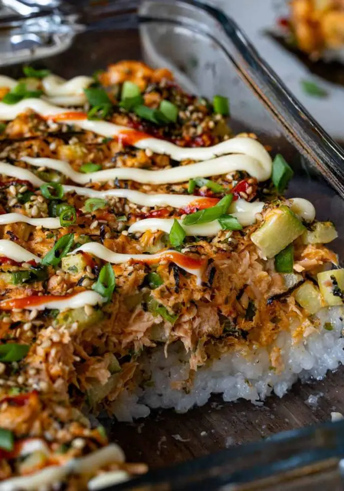 A close-up of a baked salmon sushi dish topped with sesame seeds, spring onions, spicy sauce and creamy mayonnaise, served in a glass baking dish.